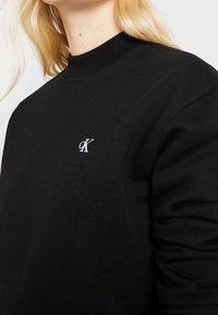 Sudadera negra con cuello acanalado, tela suave y un pequeño logo blanco bordado en el pecho. Textura suave, ajuste casual.