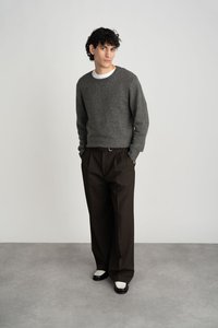 Hall tumehallist lõngast kyymaga kampsun ümmarguse kaelusega, kombineeritud tumedate pruunide volditud pükstega ja mustade kingadega, millel on valged detailid. Ühtlane taust.