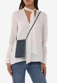 Grijze, gehaakte telefoontas voor crossbody met een dunne strap, gedragen over een witte blouse met een V-hals en een gestrikt strikdetail.