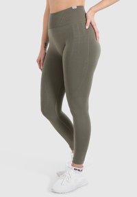 Leggings a vita alta di colore verde oliva realizzati in materiale elasticizzato; presentano una superficie liscia e dettagli delle cuciture sui lati. Abbinati a sneakers bianche.