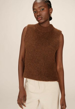 Pull en rib chocolat sans manches avec un col montant. Tissu doux et texturé ; porté avec un pantalon en velours côtelé clair et taille haute.