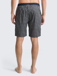Mand står barfodet med ryggen til, iført shorts i marineblå og hvide tern med elastisk talje mod en ensfarvet baggrund.