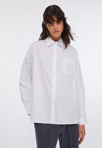 Chemise blanche à boutons avec un motif vertical subtil d'accents argentés et une poche poitrine unique, manches longues et coupe décontractée.