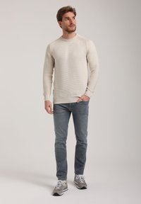 Helles beige, gestricktes Sweatshirt mit strukturiertem Muster, kombiniert mit hellgrauen Jeans und grauen Sneakers. Das Model steht mit neutralem Ausdruck.