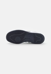 Sula av svart och vit New Balance-sneaker som visar mönstret på sulan och varumärkeslogotypen på en enfärgad vit bakgrund.