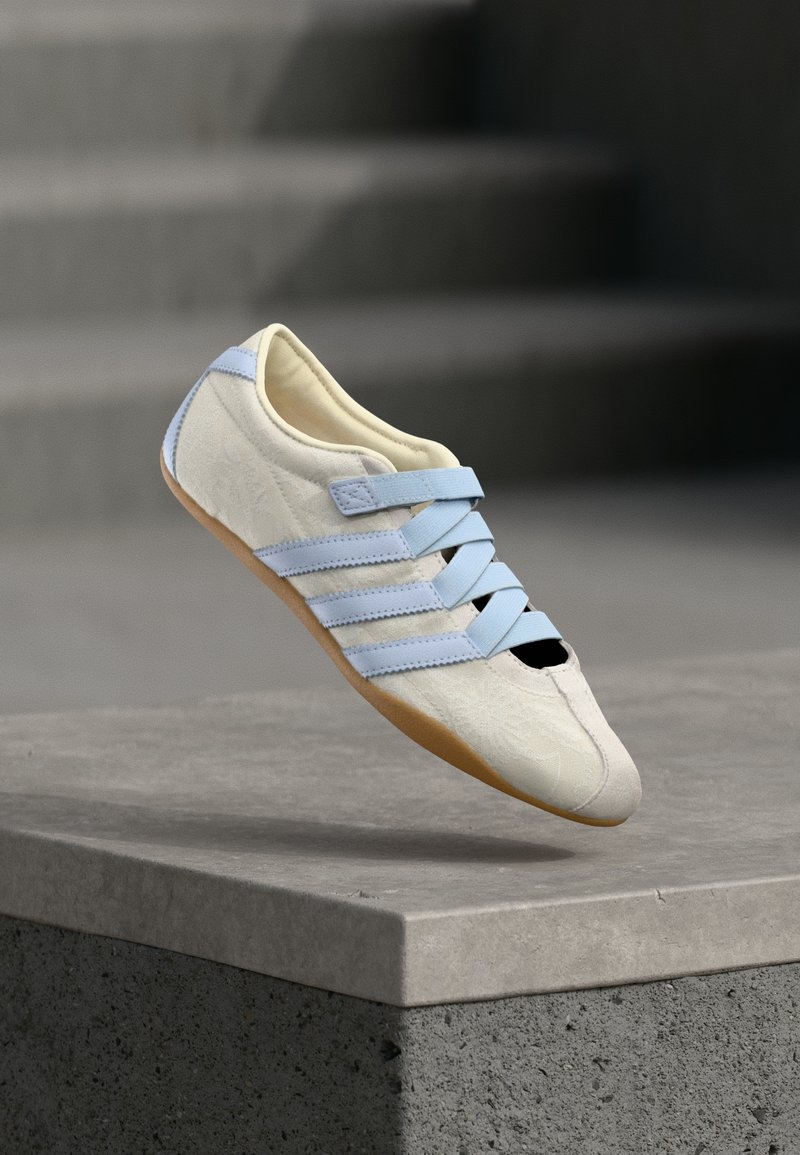 Chaussure en daim blanc avec des sangles bleu clair, un bout noir et une semelle en caoutchouc. Présente un motif floral en relief sur la tige.