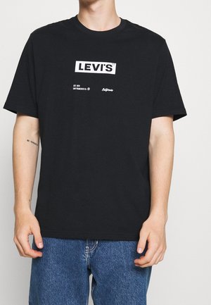 Osoba nosząca czarną koszulkę Levi's z białym logo, w zestawieniu z niebieskimi dżinsami, stojąca na jednolitym tle.