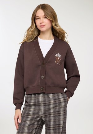 Cardigan marrone corto con scollo a V, bottoni sul davanti ed emblema ricamato. Abbinato a pantaloni grigi a quadri con una vestibilità comoda.
