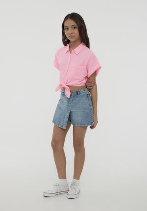 Camicia rosa a maniche corte con bottoni legata in vita, abbinata a pantaloncini di jeans azzurri a vita alta. Sneakers bianche completano l'outfit.