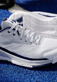 Bílé sportovní boty s síťovaným svrškem a námořnickými akcenty, s texturovanou podrážkou a symetrickým designem. Viditelné tkaničky a detaily loga.