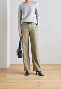 Henkilö, jolla on päällään vaaleanharmaa pitkähihaisen yläosan, beige korkeavyötäröiset housut, musta vyö ja mustat korkokengät, pitää mustaa laukkua yhdellä kädellä.