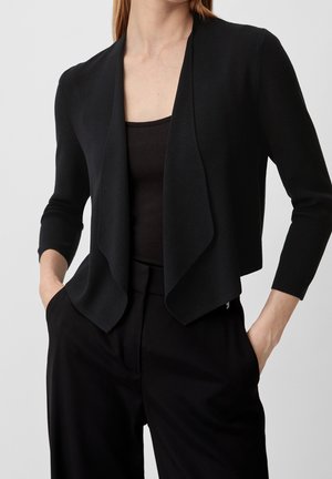 Mujer que lleva un cárdigan negro abierto por delante con mangas tres cuartos sobre una camiseta negra y pantalones negros, con las manos en los bolsillos.