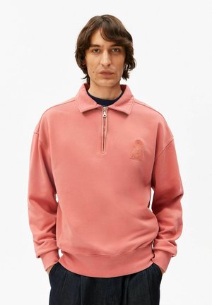 Homme portant un pull-over rose saumon avec un col demi-fermeture éclair et un logo "A" en relief sur la poitrine, les mains dans les poches d'un pantalon foncé.