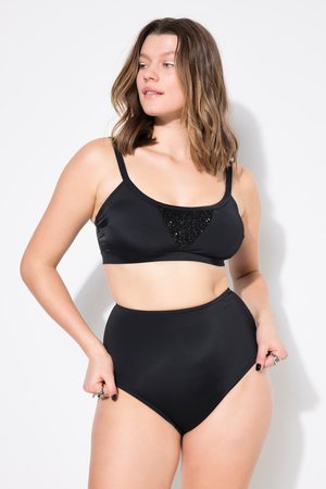 Frau trägt schwarzen Bikini mit hochgeschnittener Hose und passendem Oberteil mit Pailletten, steht vor weißem Hintergrund und schaut nach links.