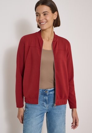 Femme souriante portant une veste rouge zippée par-dessus un haut beige et un jean bleu clair sur un fond uni clair.