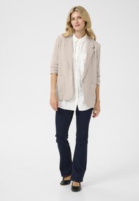 Beige blazer met twee voorzakken over een wit shirt, gecombineerd met donkerblauwe flared jeans en zwarte ballerina's. Glad stof, perfecte pasvorm.