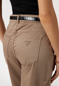 Pantalons ajustés beiges avec une texture lisse, dotés de poches arrière et d'un logo Guess en forme de triangle. Associés à une ceinture noire brillante.