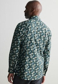 Bloemmotief shirt in donker teal met witte bloemen en groene bladeren, voorzien van een standaardkraag en lange mouwen, gemaakt van lichtgewicht stof.