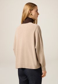 Beżowy sweter z dzianiny z brązowym golfem, z opuszczanymi ramionami i prążkowanymi detalami. Tył ma luźny krój.