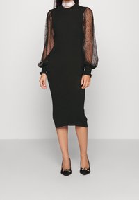 Robe noire midi avec de longues manches bouffantes en tulle transparent, dotées d'une texture à pois. Silhouette ajustée et matériau en maille côtelée. Associée à des talons noirs.