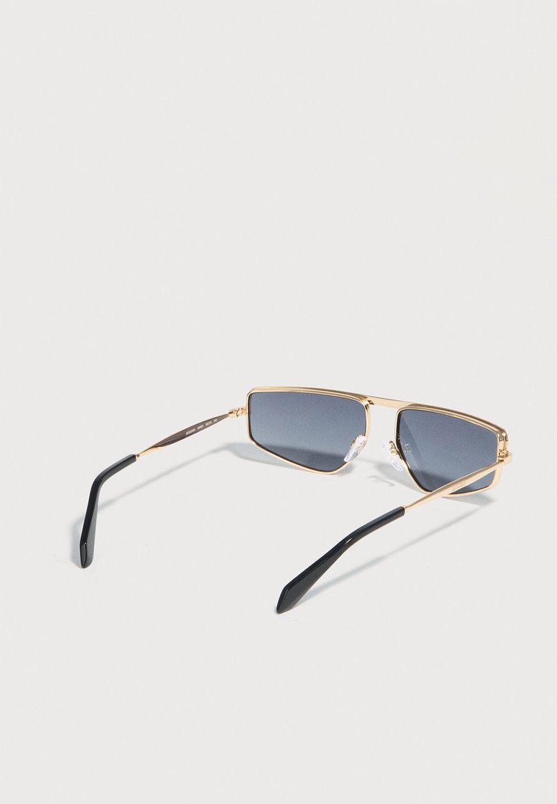 Lunettes de soleil rectangulaires à monture dorée avec des verres teintés foncés et des branches noires, posées sur un fond clair uni.