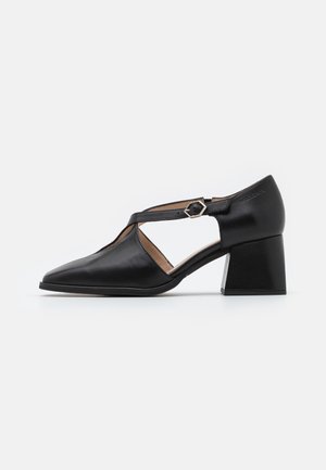 Chaussures à talons en cuir noir avec bout pointu, design ajouré, et bride à la cheville sécurisée par une boucle métallique hexagonale.