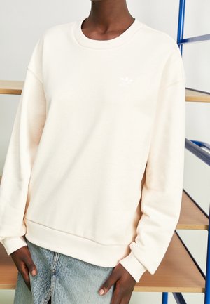 Osoba ubrana w gładką kremową bluzę z małym białym logo na piersi oraz jasnoniebieskie dżinsy, stojąca przy drewnianych półkach z niebieską ramą.