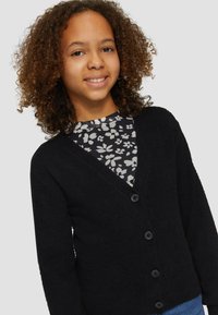 Schwarze Strickjacke aus weichem, strukturiertem Material, mit V-Ausschnitt und sechs schwarzen Knöpfen. Getragen über einem floral gemusterten Oberteil.