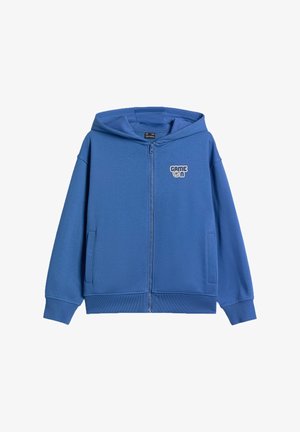 Garçon portant une casquette blanche, un sweat à capuche bleu zippé, un t-shirt blanc, un pantalon cargo foncé et des baskets gris et blanc, debout les mains dans les poches.