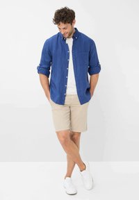 Blauwe overhemdd, opgerolde mouwen, met een patch zak, gecombineerd met beige shorts. Witte sneakers maken de casual look compleet.