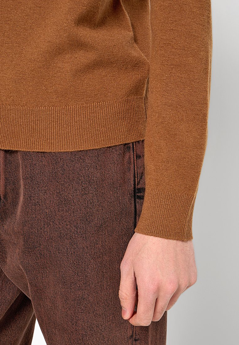 Brauner Strickpullover mit eng anliegendem geripptem Bündchen, kombiniert mit strukturierten rostfarbenen Hosen, die eine Nahaufnahme des Ärmels und der Hand zeigen.