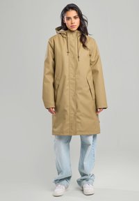 Beige waterproof parka, hooded with drawstrings, snap buttons, and side pockets. Worn over light blue baggy jeans and white sneakers.