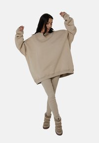 Beżowy oversize'owy sweatshirt wykonany z miękkiej tkaniny, z opadającymi ramionami i ściągaczami, zestawiony z dopasowanymi beżowymi legginsami i beżowymi botkami.