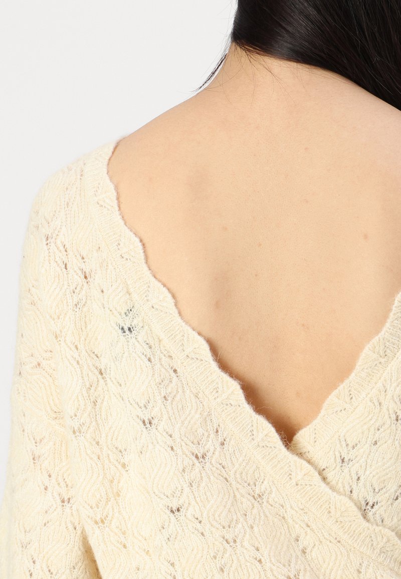 Maglione lavorato a maglia color crema con scollatura a V, caratterizzato da un motivo ondulato e una finitura testurizzata, realizzato in un materiale morbido e leggero.
