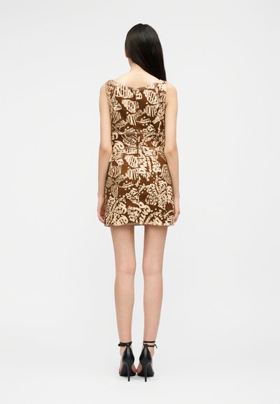 Γυναίκα με μακριά σκούρα μαλλιά που φοράει αμάνικο καφέ μίνι φόρεμα με μπεζ γκράφιτι κείμενο και μαύρα ψηλοτάκουνα πέδιλα με λουράκι στον αστράγαλο, στέκεται με την πλάτη προς τα εμπρός.