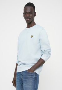 Sweat-shirt bleu clair avec poignets et ourlet côtelés, arborant un petit logo jaune sur la poitrine gauche. Assorti à un jean bleu.