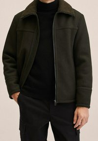 Giacca verde scuro con colletto peloso, tasche frontali e chiusura con zip. Indossata sopra un dolcevita nero, caratterizzata da un design liscio e strutturato.