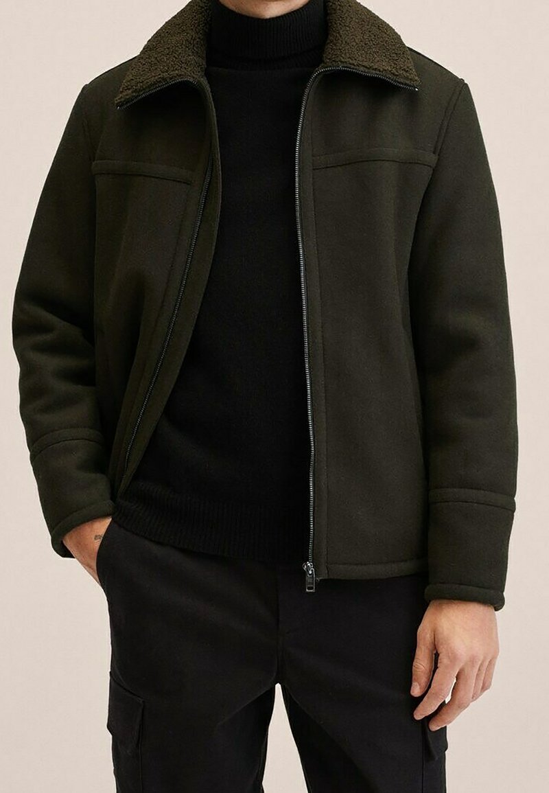 Giacca verde scuro con colletto peloso, tasche frontali e chiusura con zip. Indossata sopra un dolcevita nero, caratterizzata da un design liscio e strutturato.