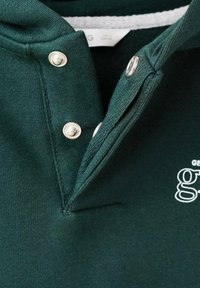 Donkergroene pullover met een kraag, voorzien van twee zilveren drukknoopjes en een witte logo-detail op de borst. Zachte, gestructureerde stof.