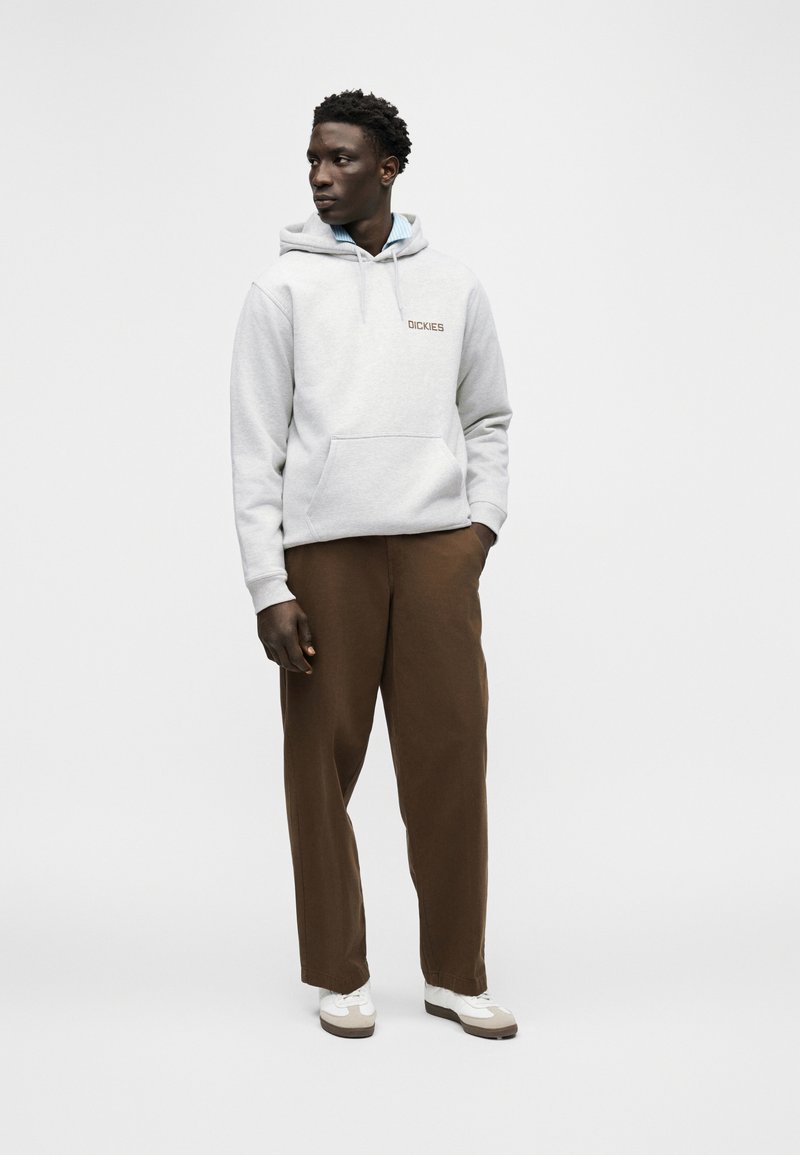 Uomo con felpa grigia chiara, maglietta blu, pantaloni marroni e scarpe da ginnastica bianche, in piedi con una mano in tasca, che guarda a sinistra.