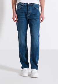 Jeans in denim blu con una linea dritta, dotati di cinque tasche e una classica chiusura a bottoni. Indossati con sneakers bianche.