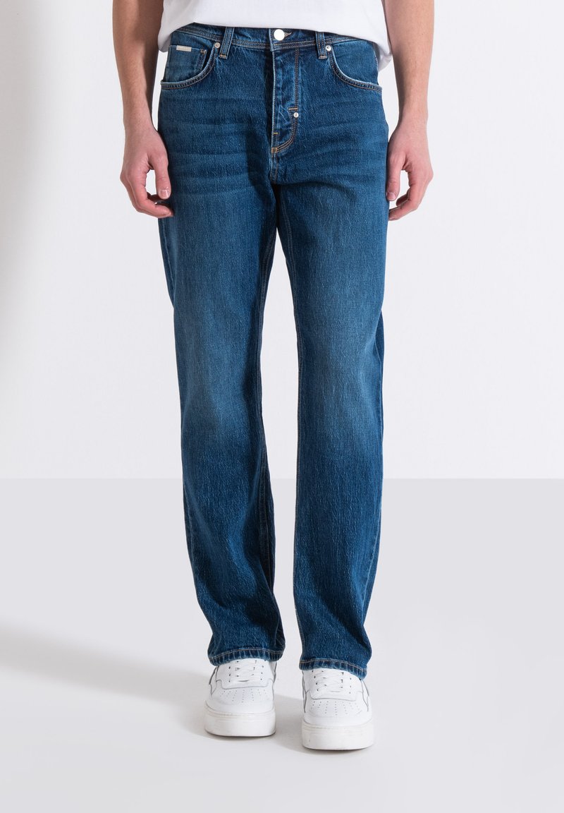 Jeans in denim blu con una linea dritta, dotati di cinque tasche e una classica chiusura a bottoni. Indossati con sneakers bianche.