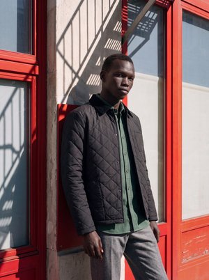 Jeune homme portant une veste matelassée noire et une chemise verte, appuyé contre un mur beige avec des fenêtres à cadres rouges au soleil.