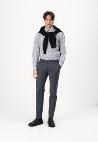 Chemise à motif floral bleu clair, pantalon gris sur mesure et chaussures noires. Un pull noir drapé sur les épaules ajoute une touche de détail.