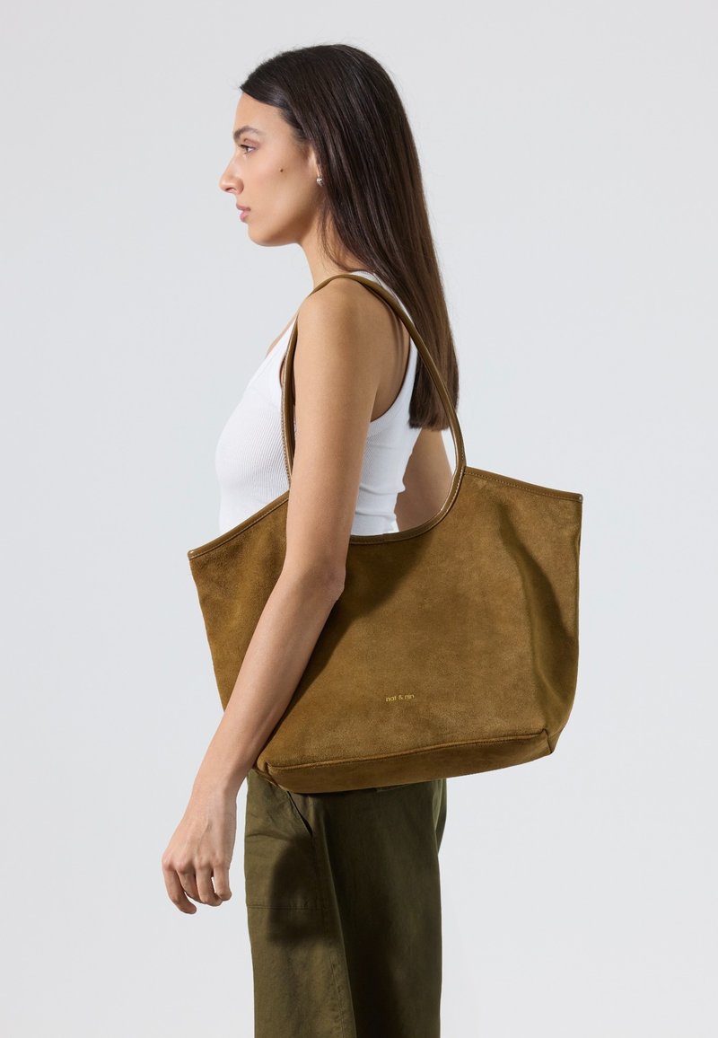 Femme en débardeur blanc et pantalon olive, portant un grand sac fourre-tout en daim marron sur l'épaule, sur fond uni.