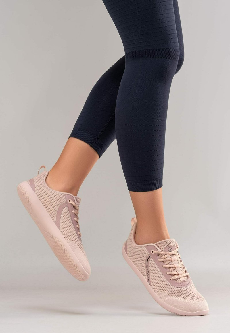 Personne portant des baskets en tricot rose clair et des leggings bleu marine foncé, pieds légèrement surélevés sur un fond gris clair uni.
