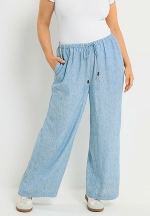 Kvinde iført lyseblå linnedbukser med vide ben, elastisk talje og snøre, kombineret med hvide sneakers og en hvid top med korte ærmer.
