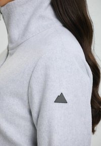 Light gray fleece jacket with a high collar and a black triangular logo patch on the left sleeve. Soft texture and fitted design.