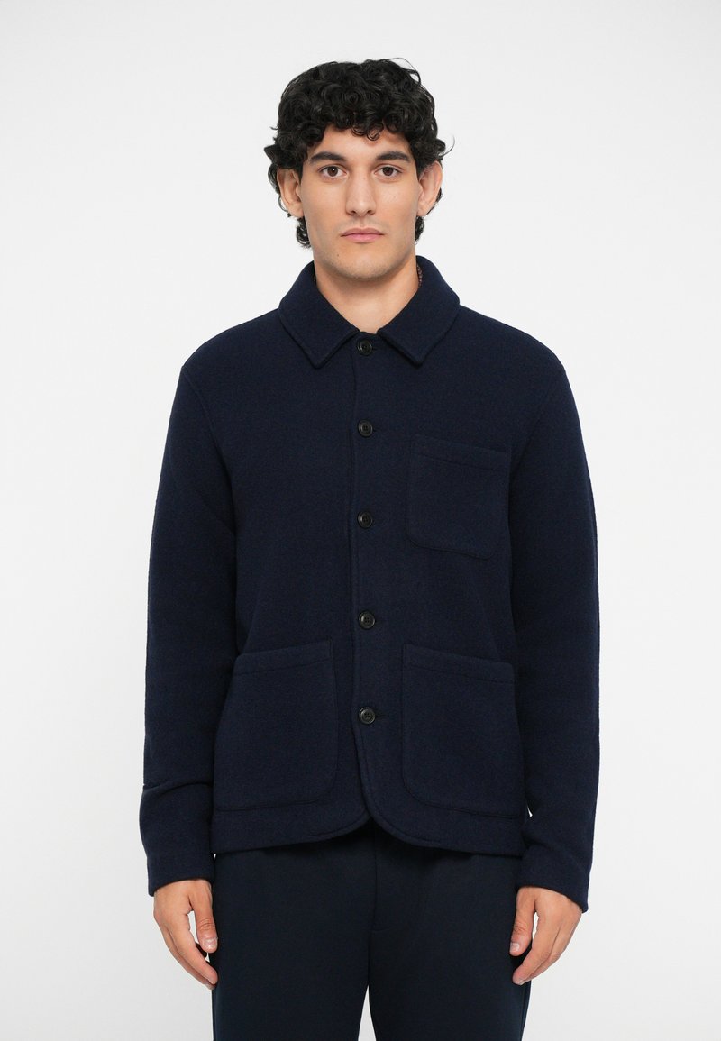 Veste en polaire bleu marine avec un col, fermeture par boutons sur le devant, deux poches avant et une coupe décontractée. Texture douce et design minimaliste.