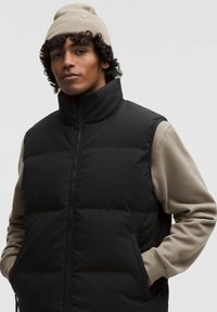 Schwarze Pufferweste mit hohem Kragen, Reißverschluss und beiger Langarmbluse darunter. Sanfte Textur, minimalistisches Design.